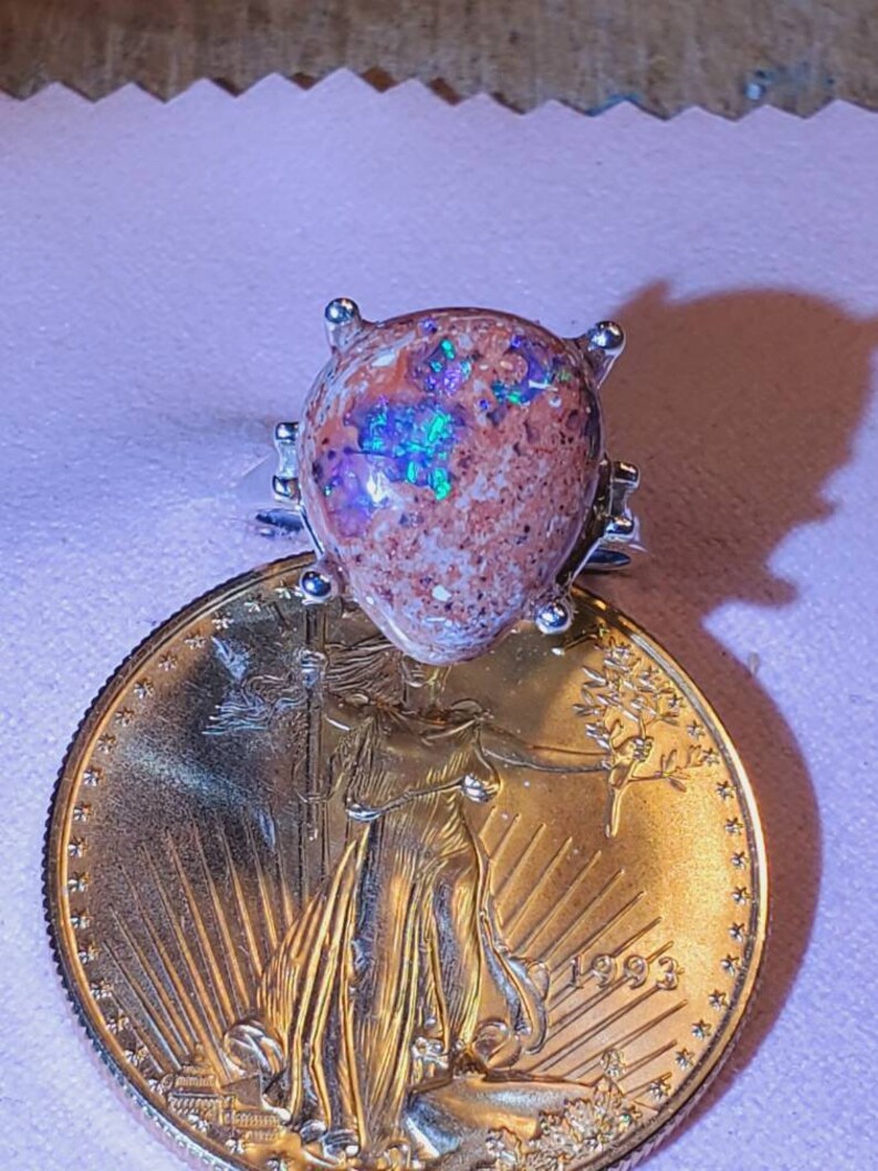May include: A silver ring with a large, pear-shaped opal gemstone. The opal is a mix of brown, white, and green with iridescent flashes of color. The ring is sitting on top of a gold American Eagle coin.