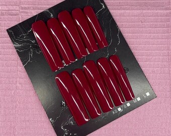 May include: A set of long, coffin-shaped false nails in a deep burgundy color. The nails are arranged on a black card with a marble-like pattern. Sizes are indicated as XS, S, M, and L. The background is a pink, textured surface.