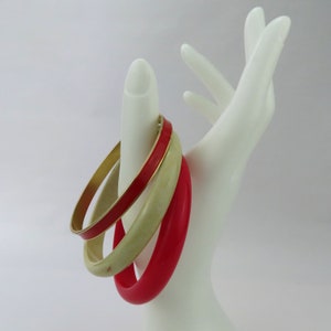 May include: Three bangles in a variety of colors. One is red with a gold rim, one is a pale green, and the other is a bright red. The bangles are displayed on a white hand-shaped jewelry holder.