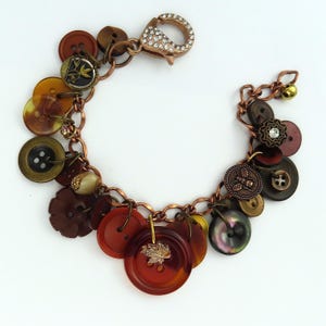 May include: A button bracelet with a copper chain and clasp. The bracelet features a variety of buttons in shades of brown, red, and yellow. Some buttons have decorative details, and a charm with a leaf is attached.