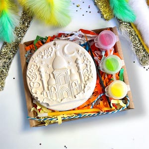 May include: A craft kit featuring a white, circular plaster relief of a mosque with a crescent moon. The kit includes three paint pots in pink, green, and yellow, along with a paintbrush and colorful shredded paper, all inside a cardboard box.