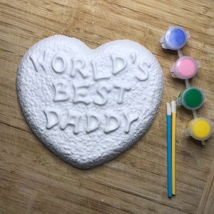 May include: White ceramic heart-shaped ornament with the words "World's Best Daddy" embossed on the front. The ornament is accompanied by four small bottles of paint in blue, pink, green, and yellow, and two paintbrushes.