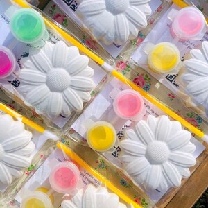 May include: A set of white ceramic daisy flower shapes with paint pots and paintbrushes for a craft project. The paint pots are in various colors, including pink, yellow, and green.