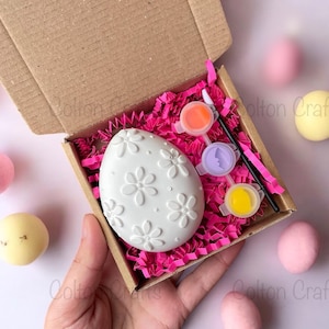 May include: A white ceramic egg with a floral design, ready to be painted. The kit includes three small pots of paint in orange, purple, and yellow, a paintbrush, and pink crinkle paper.