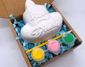 May include: A white, unpainted diya-shaped craft kit with the words "Happy Diwali" in cursive. The kit includes three paint pots in yellow, pink, and green, a paintbrush, and blue paper filler, all inside a brown cardboard box.