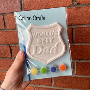 May include: A white ceramic paint-your-own plaque shaped like a shield with the words "World's Best Dad" on it. The plaque is packaged with a paintbrush and four small bottles of paint in yellow, green, blue, and orange.