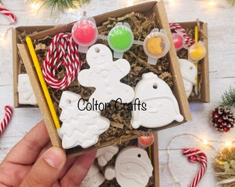 May include: A craft kit in a cardboard box containing white ceramic ornaments shaped like a gingerbread person, a Christmas tree, a snowman, and Santa Claus. Includes a yellow paintbrush, red and white twine, and small pots of paint.