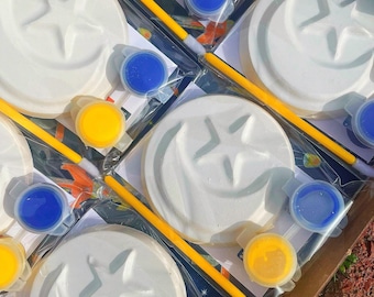 May include: A set of four white ceramic paint-your-own ornaments featuring a crescent moon and star design. Each ornament comes with a small paintbrush, yellow, blue, and white paint.