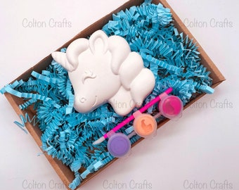 May include: A white ceramic unicorn head figurine in a brown box with blue crinkle paper. The box also contains three small bottles of paint and a pink paintbrush.