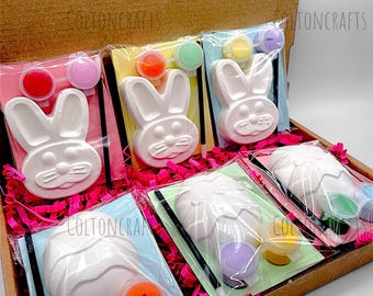 May include: A set of six Easter-themed paint-your-own crafts in a cardboard box. The crafts include bunny and egg shapes with paint pots and brushes. The box is filled with pink shredded paper.