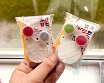 May include: Two white ceramic ornaments, one with a star and the other with the text "Merry Christmas!"  Each ornament is packaged with a small bottle of paint, a paintbrush, and a string. The ornaments are labeled "Colton Crafts Co."
