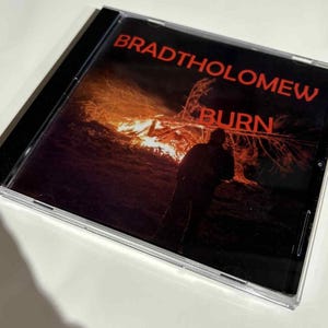 May include: A CD case featuring the album "BURN" by Bradtholomew. The cover art depicts a silhouetted figure in front of a bonfire with the album title in red. The case is clear with a black spine.