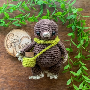 Puede incluir: Un topo de crochet marrón con una bufanda verde y una bolsa verde con una tapa marrón. El topo está sentado sobre una superficie de madera con hojas verdes en el fondo.