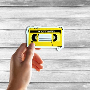 May include: Yellow VHS tape sticker with black dripping paint. The tape has a heart and the text "I &hearts; horror movies!" on the label.