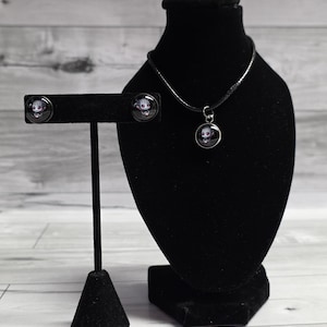 May include: A black necklace and matching stud earrings set. The jewelry features a small, round pendant and earrings with a skull design. The set is displayed on a black jewelry bust and earring stand against a light wood background.