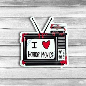 May include: A black and white sticker of a vintage television with a red heart and the text "I love horror movies". The television has red dripping blood on the sides and a stack of VHS tapes on top. The tapes are labeled "IT", "HALLOWEEN", "SAW", and "THE TEXAS CHAINSAW MASSACRE".