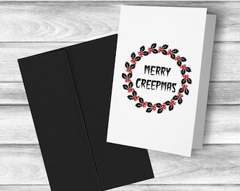 May include: A white greeting card with a black envelope. The card features a wreath of black leaves and red berries around the words "MERRY CREEPMAS" in black, gothic-style lettering. The background is a light wood grain.