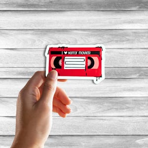 May include: Red VHS tape sticker with dripping black paint. The tape has a white label with the text "I &hearts; horror movies!" and black spools.