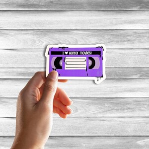 May include: A purple VHS tape with black dripping paint. The tape has the text "I &hearts; horror movies!" on it.