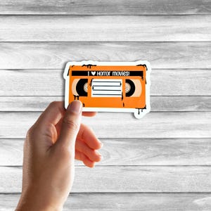 May include: Orange VHS tape sticker with black dripping paint around the edges. The tape has a black label with the text "I &hearts; Horror movies!" and a black and white image of a cassette tape.