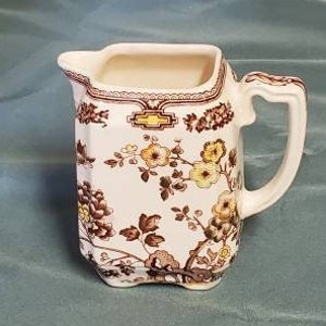 May include: A white ceramic pitcher with a brown and yellow floral pattern. The pitcher has a handle and a square base.