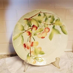 May include: A white ceramic plate with a hand-painted design of a cherry branch with red and yellow cherries. The plate is on a white crocheted doily.