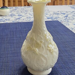May include: A cream-colored glass vase with a ruffled rim and a bulbous base. The surface features a raised, embossed pattern of swirling vines and leaves with a subtle pearlescent finish, creating a textured appearance across the entire body.