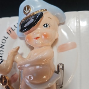 Puede incluir: Una figurita de cerámica de un bebé que lleva un gorro de marinero azul con ribete dorado y visera negra. La figurita sostiene un pequeño objeto dorado en su mano izquierda. La figurita está unida a una base de cerámica blanca con la palabra "Honolulu" escrita en ella.