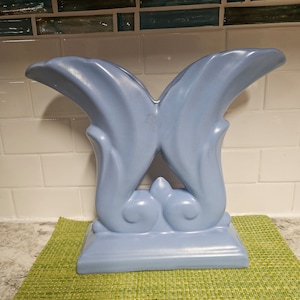May include: A light blue ceramic butterfly sculpture with a stylised design. The sculpture has a base and two wings that are curved and pointed.
