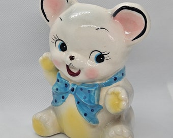 May include: A ceramic money box shaped like a white bear with a blue polka dot bow tie. The bear has pink ears, black eyes, and a yellow tummy.