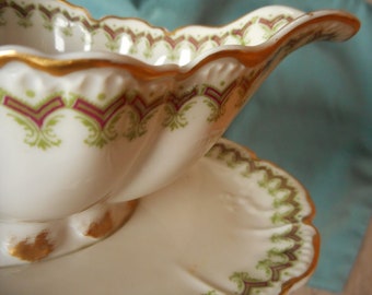May include: A white porcelain gravy boat with a gold rim and green and burgundy floral pattern. The boat is sitting on a matching saucer.