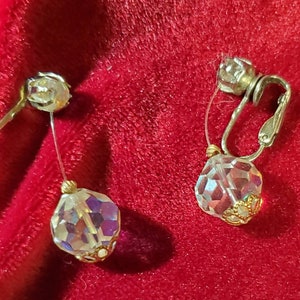 Può includere: Un paio di orecchini a clip argentati con gocce di cristallo trasparente iridescente. I cristalli sono sfaccettati e hanno un bordo dorato.