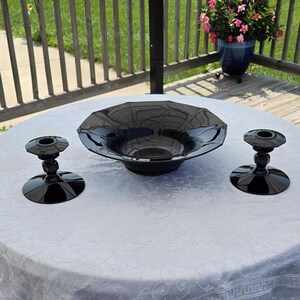 May include: A set of three black glass items consisting of one wide, shallow bowl with a faceted octagonal rim and two matching candle holders with circular bases and tiered stems, all featuring a smooth, reflective, and glossy dark finish.