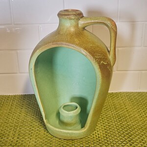 Puede incluir: Un portavelas de cerámica verde y marrón con forma de jarra con un hueco para una vela. El portavelas tiene un aspecto rústico y vintage.