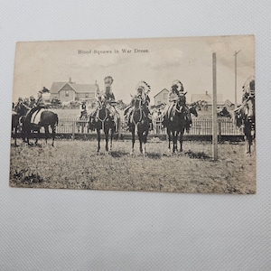 May include: Black and white vintage postcard showing a group of Native American women on horseback in war dress. The text on the postcard reads "Blood Squaws in War Dress."