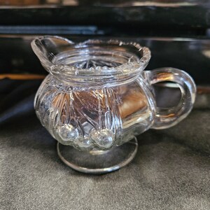 May include: A clear glass creamer with a decorative pattern of raised dots and a scalloped rim. The creamer has a handle and a round base.
