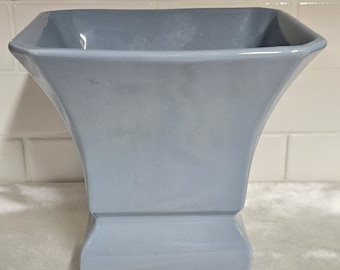 May include: A light blue ceramic vase with a square opening and a flared base. The vase is made of glazed ceramic and has a smooth finish.