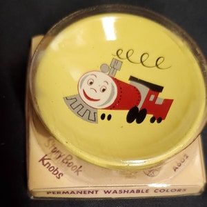 Può includere: Un pomello di plastica giallo con un disegno di un treno dei cartoni animati. Il treno è rosso con un viso grigio e ruote nere. Il pomello è etichettato "Story Book Knobs" e "Permanent Washable Colors".