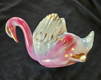 May include: A pink and white ceramic swan figurine with gold accents. The swan is in a curled position with its head turned to the side.