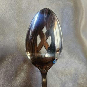 May include: A silver slotted spoon with a diamond-shaped pattern cut out of the bowl. The spoon is shiny and reflects light.