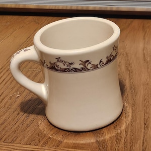 May include: Cream-colored ceramic mug with a curved handle and a brown decorative floral pattern around the rim. The mug has a slightly angled design, giving it a unique shape. The mug is sitting on a wooden surface.