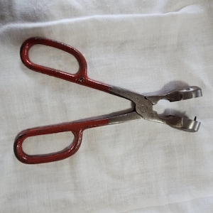 Puede incluir: Alicates de metal con mango rojo y una mandíbula plateada con la palabra "Divin" impresa en el lateral.