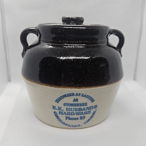 Könnte beinhalten: Eine braune und weiße Steinzeugkrug mit Deckel. Der Krug hat ein blaues und weißes Etikett mit der Aufschrift "Hardware as Lasting as Stoneware E.K. Husbands Hardware Phone 89 Gresham, Neb."