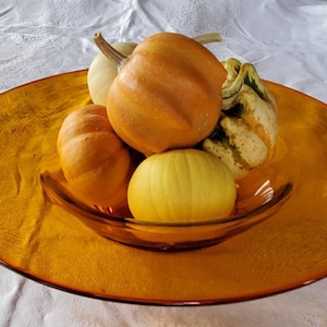 Puede incluir: Una colección de calabazas y pequeñas calabazas en varios tonos de amarillo y naranja, expuestas en un cuenco de vidrio transparente sobre un plato grande de color ámbar.