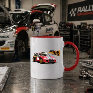 Può includere: Tazza in ceramica bianca con interno e manico rossi, con grafica di auto da rally rossa con la scritta "SPECIAL STAGE". Lo sfondo mostra un'auto in un'officina, con attrezzi e attrezzature.
