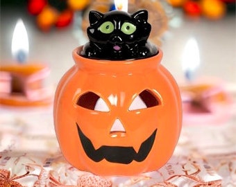 May include: A ceramic Halloween candle holder shaped like an orange jack-o'-lantern with a carved face. A black cat figurine sits atop the pumpkin, and a lit candle is inside. The background is blurred, with other lit candles visible.