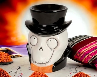 May include: A ceramic skull-shaped mug wearing a black top hat and tuxedo. The mug has a smiling face with large eyes and a tan collar. The background is a blurred orange and yellow.