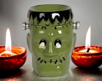 May include: A green ceramic Frankenstein's monster head-shaped candle holder with black hair and silver bolts on the sides. Two lit candles in red holders flank the monster's head, creating a spooky Halloween display.