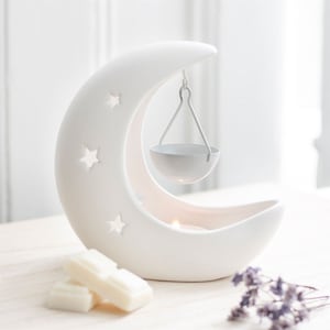 May include: White ceramic crescent moon wax melt warmer with a metal hanging bowl and a small tea light candle. The warmer has cut-out stars and is on a white surface with wax melts and dried lavender.