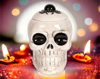 May include: White ceramic skull shaped jar with a black spider on the lid. The jar has black eyes and a toothy grin.